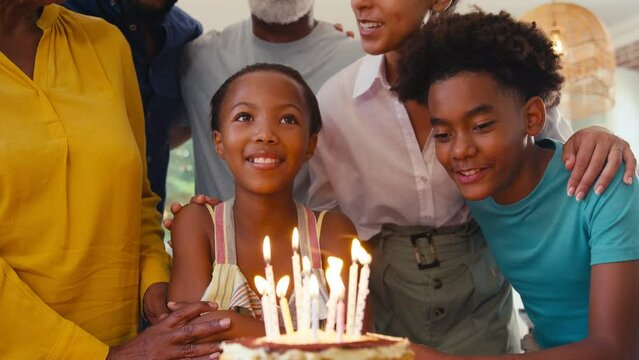 Close up of loving multi-generation family standing in kitchen at home celebrating granddaughter's birthday with cake and candles singing happy birthday to her as she blows out candles - shot in slow 