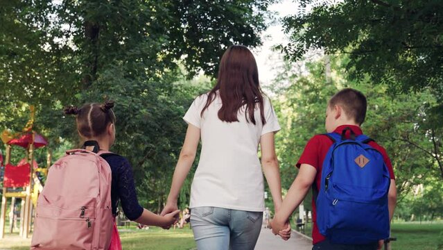 Happy family mom daughter son holding hands go to school on path. People with backpacks walking in park. School children with their mother go to school. Boy girl mom walking outdoors. Concept family.