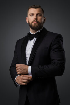 Confident and stylish handsome man with a manicured beard wears a luxurious black suit and bowtie on a grey background