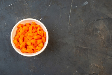 Above view of chopped fresh carrots in a white bowl on the right side on dark color table with free space