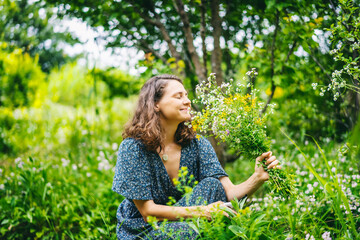 Young cheerful smiling woman in a summer dress picking a bouquet of wild flowers in a summer garden