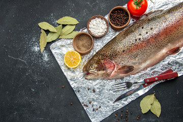 Above view of scales removed fresh fish cutlery set bay leaves tomato and different spices on dark color background