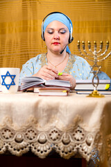 A Jewish woman in a kisui rosh headdress, a Torah teacher in headphones with a microphone, reads...