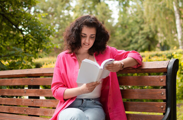 Focused young girl in bright clothes is reading book and sitting on park bench. Concept of education. Summer concept. Self-confident.