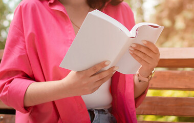 Focused young girl in bright clothes is reading book and sitting on park bench. Concept of...