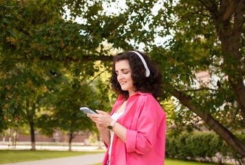 Fototapeta premium Woman in profile in bright clothes with headphones listens to music, breathes fresh air, relaxing in the park. Concept of urban lifestyle of people. Summer concept. Enjoys life, gets pleasure