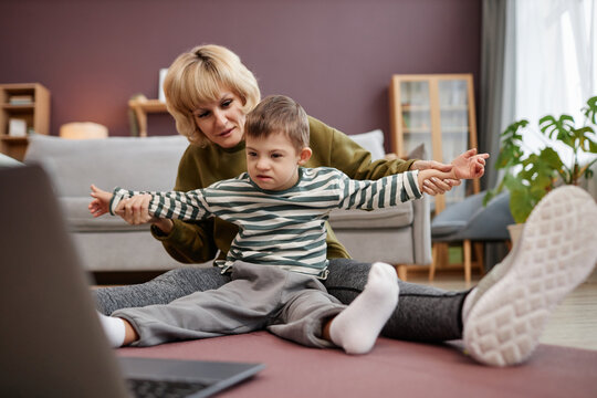 Full length portrait of mother and son with down syndrome watching videos via laptop together while sitting on floor at home