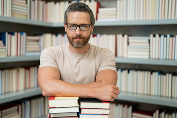 Portrait of teacher with book in library classroom. Handsome teacher in university library. Teachers Day. Teacher giving classes. School teacher in library. Tutor at college library on bookshelf.