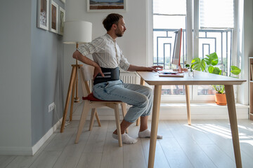 Man in sick leave works types on computer keyboard at home wearing back support belt corset on lower back during exacerbation to treatment of hernia. Back pain as consequences of sedentary work. 