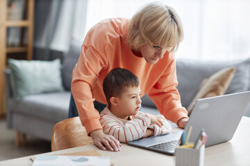 Portrait of little boy with down syndrome studying at home and using computer with mom assisting,...