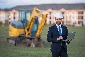 Architect at a construction site. Architect man in helmet and suit at modern home building construction. Architect with a safety vest and suit. Confident architect standing at house background.