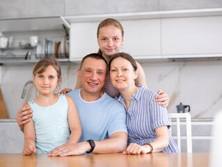 Obraz na płótnie Canvas Happy family of four people is sitting at desk. Smiling parents with daughters sit on one side of kitchen table and look at camera