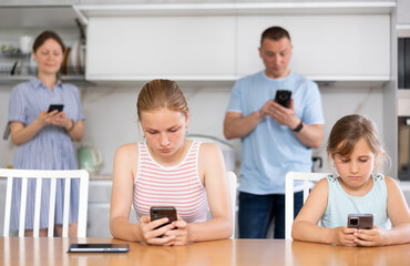 Fototapeta premium Sister girls sitting near kitchen table with phones in hands and flipping through tape and liking photos of friends, parents surf Internet in background