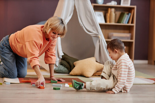 Side view portrait of little boy with down syndrome sitting on floor in playroom and playing with toy blocks mom helping