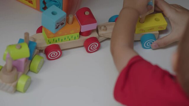 educational toys baby development activities with mother home daytime. mom together toddler kid play colorful wooden train on white table. baby boy red shirt female hand help build parts. motor skill