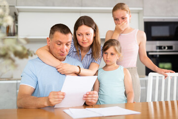 Obraz na płótnie Canvas Saddened father reads studies financial expenditure documents. Thoughtful man is encouraged and hugged by wife and daughters. Woman with husband carefully reads sad news in financial business report