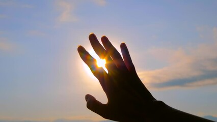 Woman silhouette is waving her hand against the sunset, sunrise sky, sun is shining through fingers - sun lens flares, close up. Calm, freedom, summer and inspiration concept
