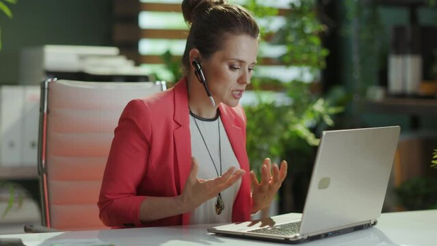 Sustainable workplace. angry modern business woman in modern green office in a red jacket with headset and laptop.
