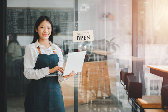 Beautiful young barista woman in apron holding laptop and standing in front of the door of cafe with open sign board. Business owner startup concept.