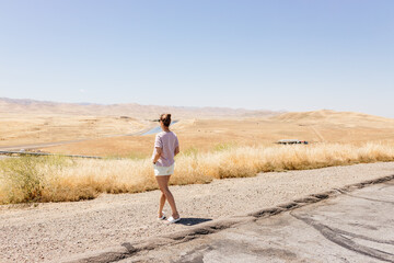A beautiful young girl stands on a hill and looks at the beautiful California summer landscape. Summer landscape with dry yellow grass and highway aerial view