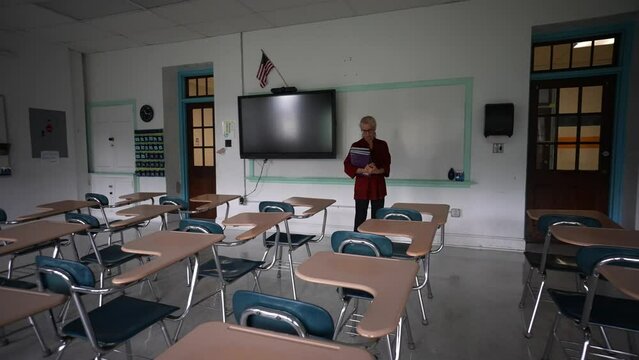 Wide angle push in view of sad teacher in an empty classroom with the lights turned off during a pandemic wondering about the future of education.