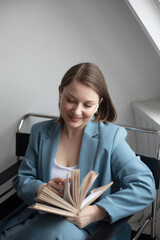 relaxed trendy middle aged woman withlight brown hair sitting in modern chair with book near window