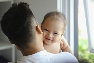 Cute Asian baby smiling on father’s shoulder near the window in the living room