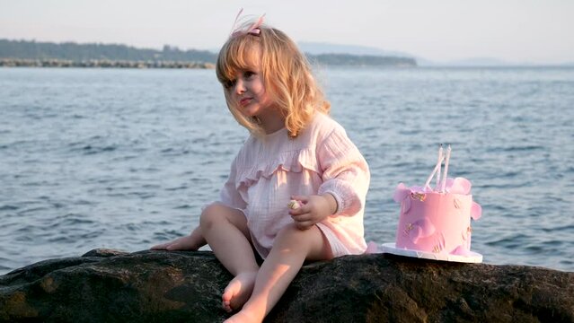 girl demands something screaming pulls hand waving fingers 3 years old child whims knocking hand on stone. angry cake pink dress candles birthday aggression spoiled child against backdrop of ocean