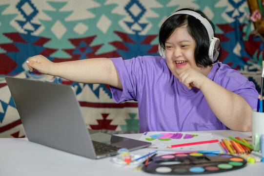 Asian girl with down syndrome wearing casual clothes using laptop at home she is very happy and excited doing winner pose with arms up smiling and screaming for success