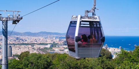 The funicular passes the support tower with a view from above of Barcelona and the sights. A trip by cable car to the observation decks. The funicular moves along the cable against the blue sky. - obrazy, fototapety, plakaty
