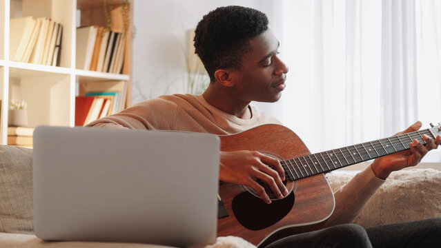 Online music school. Guitar teacher. Remote learning. Man teaching string instrument lesson class on laptop internet video call on couch at home.