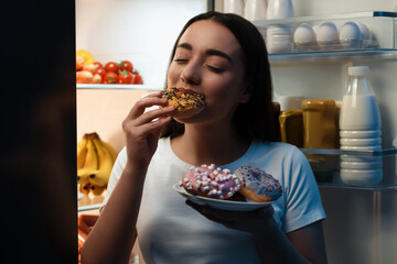 Young woman eating donut near refrigerator in kitchen at night. Bad habit