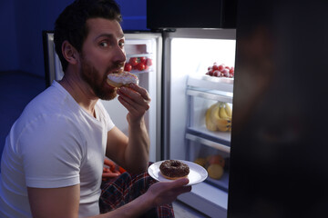 Man eating donuts near refrigerator in kitchen at night. Bad habit