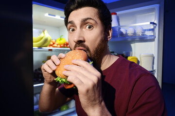 Man eating burger near refrigerator in kitchen at night. Bad habit