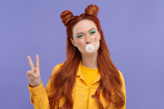 Portrait of beautiful woman with bright makeup blowing bubble gum and showing peace gesture on violet background