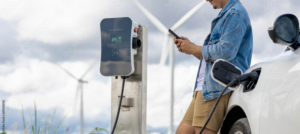 Sticker Progressive man with his electric car, EV car recharging energy from charging station on green field with wind turbine as concept of future sustainable energy. Electric vehicle with energy generator.