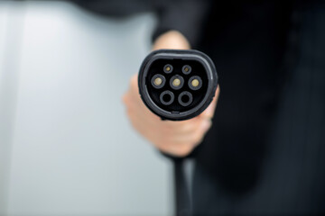 Focus closeup hand holding EV plug for electric vehicle pointing at camera with charging station at...