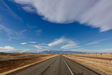 Landscape with a highway among meadows and trees in the foreground, blue mountains with snow in the background and a bright blue sky with large white clouds. Scenic landscape on a sunny autumn day