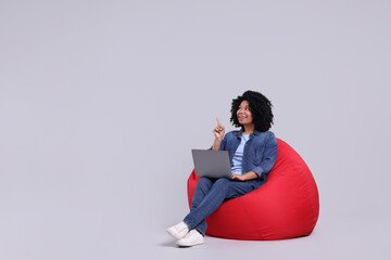 Happy young woman with laptop pointing at something on beanbag chair against light grey background. Space for text