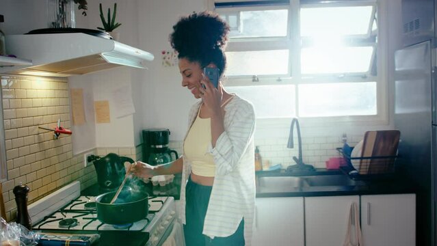 Young black woman stirring boiling food in pot on gas stove and speaking on mobile phone, cooking dinner in the kitchen at home. Medium long shot