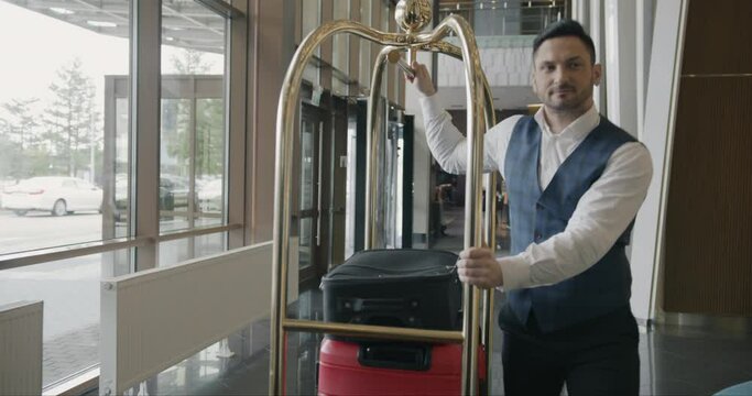 Dolly shot of helpful bellman pushing trolley with luggage along hotel corridor working alone wearing uniform. People and occupation concept.