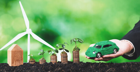 Businessman holding EV car model with seedling grow on coin stack and wind turbine. Eco-friendly...