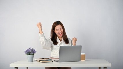 A cheerful Asian businesswoman sits at her desk, raising her hands, screaming with joy