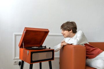 Handsome teenager boy listening vinyl records on retro wooden turntable, laying on brown sofa and...