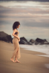 This captivating photograph of a stunning 30 year-old young, slim, and athletic caucasian model with curly hair  posing at the beach. Dressed in a beige crochet beach dress.