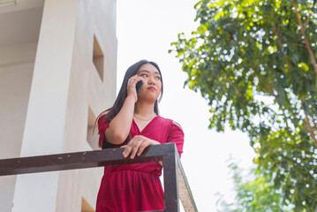 A white Asian lady in a long red dress holding on a rail, gazing far away with a fierce face while talking to someone on the phone. A tree and building is shown in the background.