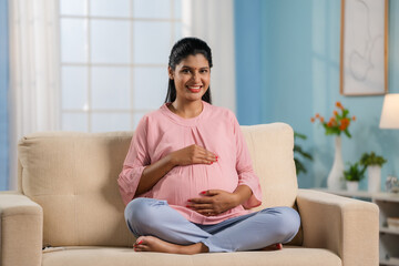 Happy indian pregnant woman feeling baby by looking and holding her tummy at home - concept of...