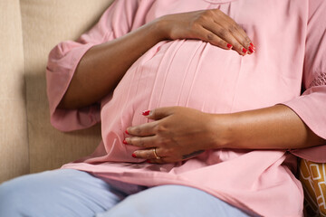Fototapeta na wymiar Close up shot of indian pregnant woman feeling by touching tummy while sitting on sofa at home - concept of motherhood, expecting and prenatal bonding