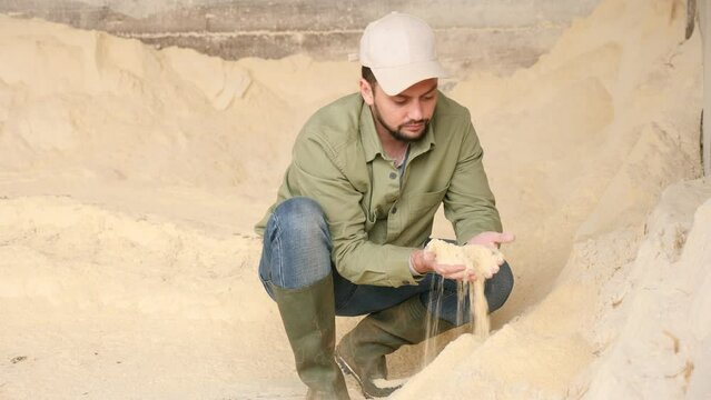 Male farmer squatting at heap of corn flour and holding bunch of it in hands.