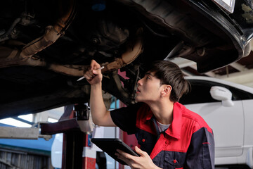 Professional young Asian male motor mechanic inspects undercarriage of electric car(EV) lifted by forklift jack for repair at garage, automotive maintenance service works industry occupation business.